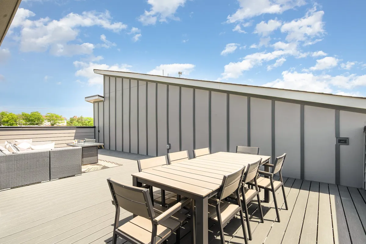 A spacious rooftop patio featuring a large dining table ideal for group meals, complemented by a sectional seating area and open sky views. The arrangement of the furniture suggests comfort and sociability, while the elevated perspective hints at expansive city vistas beyond the patio edge.