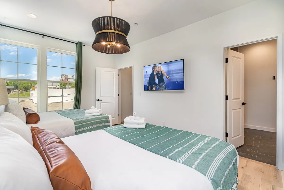 A bright, cheerful bedroom with beds dressed in striped blankets, large windows letting in natural light, a television showing a lively scene, and a modern light fixture. The space looks both cozy and functional.