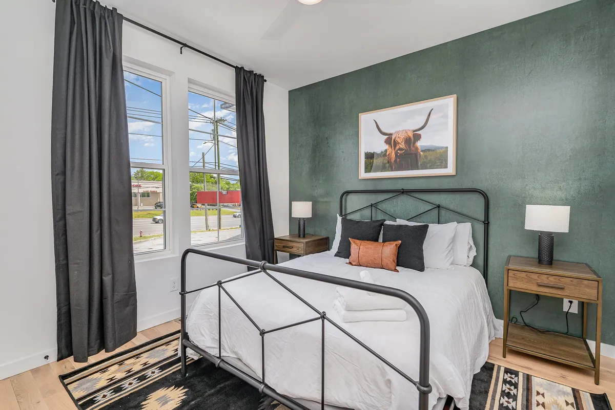This bedroom features a single bed with crisp white linens, a bold green accent wall, large windows letting in daylight, and distinctive artwork of a bull. The setting is relaxed and modern, with an emphasis on personal retreat and style.