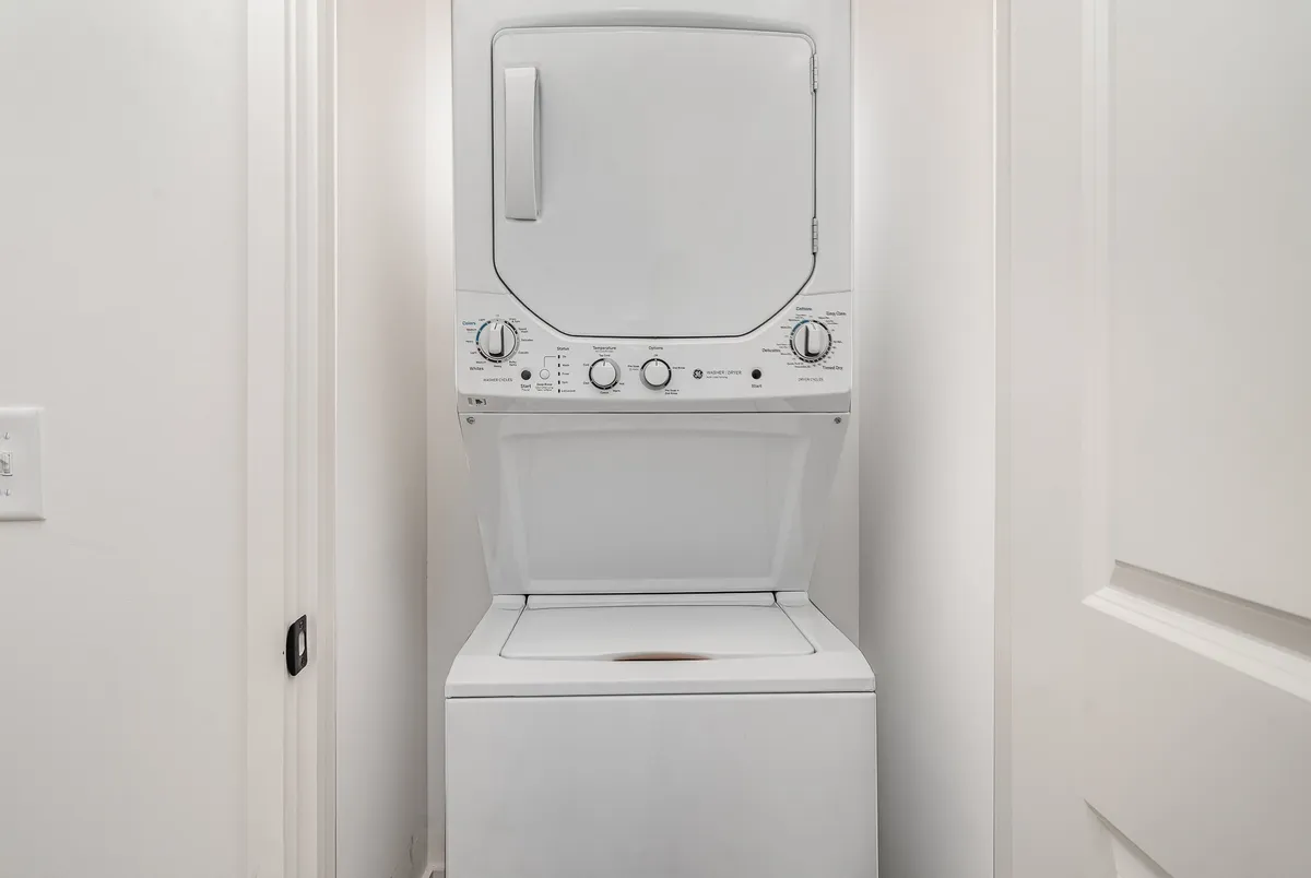 This image depicts a clean, modern laundry area featuring a stacked washer and dryer set against crisp white walls and a simple door frame. The space looks organized and practical, perfect for guests who value convenience during their stay.