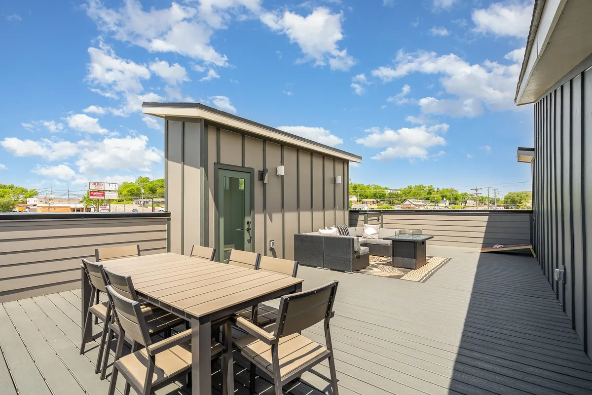 A stylish outdoor dining area with a large table, cushioned seating, and a distinctive green door. The space is set for social gatherings, offering a cozy spot for sunset dinners or morning coffee, with hints of skyline views in the background.