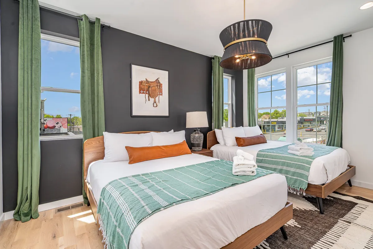 The image displays a primary bedroom with two beds adorned with decorative throws, large windows allowing natural light to fill the room, green curtains, stylish pendant lighting, and tasteful wall art. The twin beds and ample space capture a sense of comfort and practicality ideal for groups or families.
