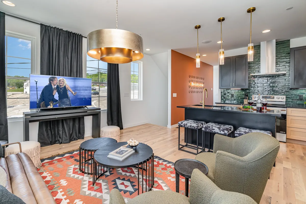This image presents a lively, open-concept living space featuring a modern seating arrangement, a television displaying a performance, a colorful area rug, pendant lighting, and a kitchen island visible in the background. The setup suggests a seamless flow between living and kitchen areas, ideal for socializing and group gatherings.