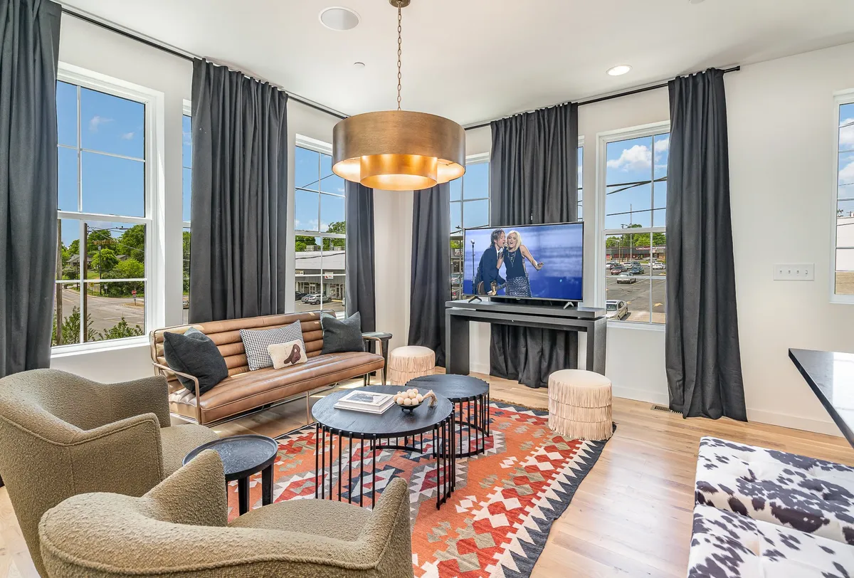 The image showcases a sun-drenched living area with expansive windows, a contemporary chandelier, comfortable sofas arranged for conversation, a plush area rug, and a television displaying inviting imagery. The space radiates warmth and modern style, perfect for gathering with friends or family.