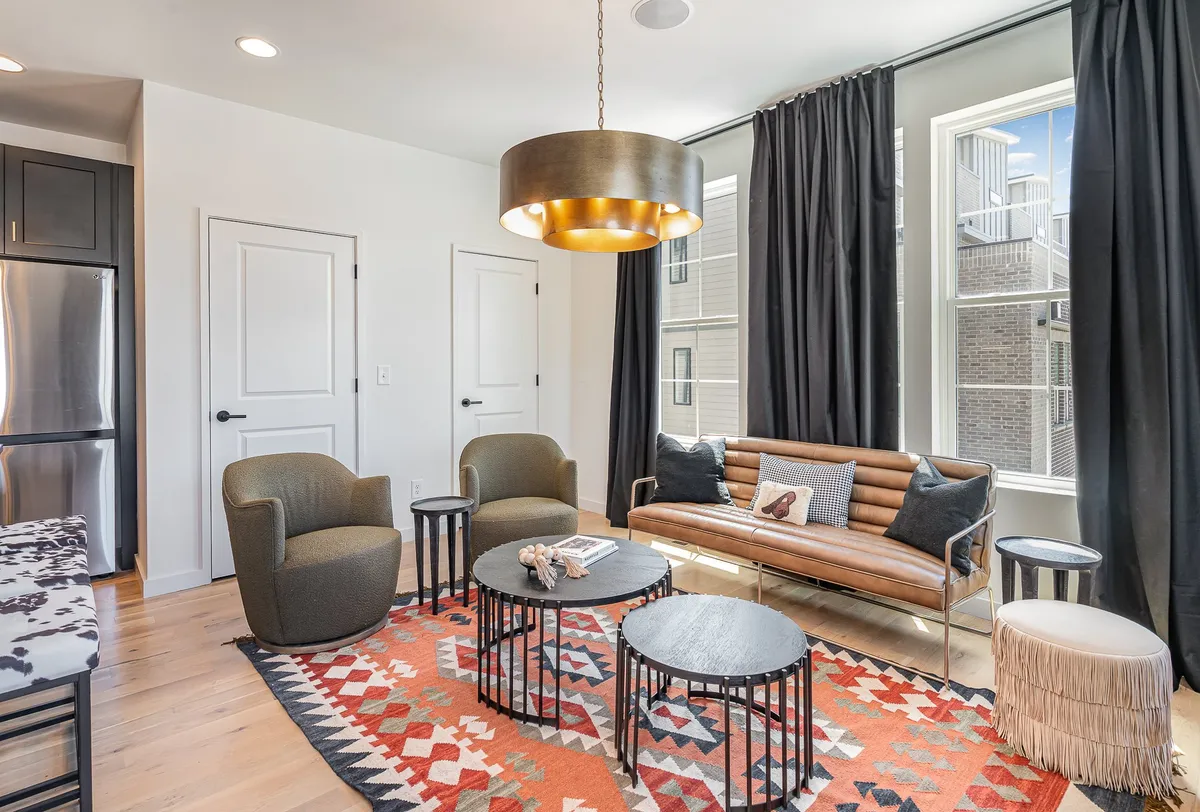 The photo shows a living area with a plush sofa, chairs, a patterned rug, and a stylish light fixture. The room feels inviting and is designed for relaxation and socializing, with natural light and comfortable seating that supports a welcoming home atmosphere.
